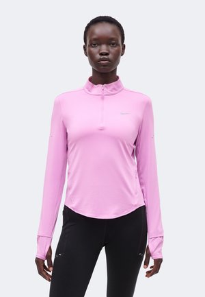 Frau trägt ein hellrosa langärmeliges Sportoberteil mit halbem Reißverschluss und schwarze Leggings, steht vor einem einfachen hellen Hintergrund.