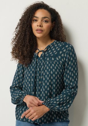 Femme aux cheveux bouclés portant un chemisier vert foncé à motifs avec des manches longues et un détail noué près de l'encolure.