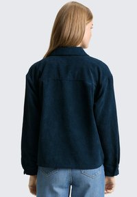 Marineblauwe corduroy jas met een kraaghals, zichtbare ribbeltextuur en knoopsluitingen bij de mouwen. De achterkant toont een recht gesneden model en yoke-detail.