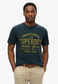 Mørkeblå bomulds T-shirt med rund halsudskæring og korte ærmer, med fedt gult tekst "SUPERDRY" og "WORK-WEAR" i et stort skriftmønster.
