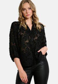 Black sheer blouse featuring textured floral patterns, a collared neckline, and button closure. Styled with fitted black pants.