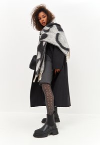 Manteau noir sur un pull gris, associé à une jupe imprimée jusqu'à mi-cuisse et des bottines noires texturées. Enveloppé dans une écharpe grise et noire.