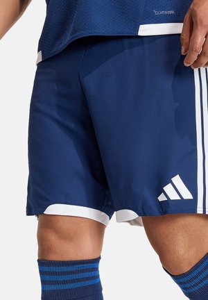 Mężczyzna ubrany w niebieskie sportowe spodenki Adidas z białymi paskami i logo Adidas, w połączeniu z niebieskimi skarpetkami w paski.