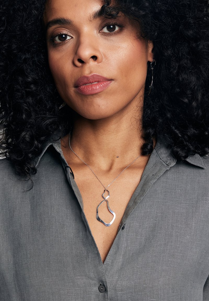 Kobieta z kręconymi czarnymi włosami, ubrana w szary koszulowy T-shirt i srebrny abstrakcyjny naszyjnik, patrzy bezpośrednio na widza.