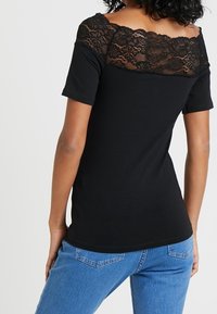 Camisa negra de manga corta con un diseño de encaje en la parte superior de la espalda. Tejido suave, corte ajustado y dobladillo redondeado. Jeans visibles por debajo.