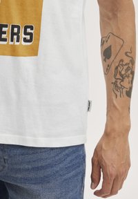 Valge puuvillane T-särk, millel on julge musta ja kuldse trükiga disain. Lühikesed varrukad, sirge äärega ja väike brändi silt küljel.