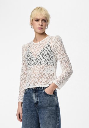 White lace long-sleeve top featuring floral patterns, a round neckline, and a cropped hem, layered over a black bralette and paired with denim jeans.