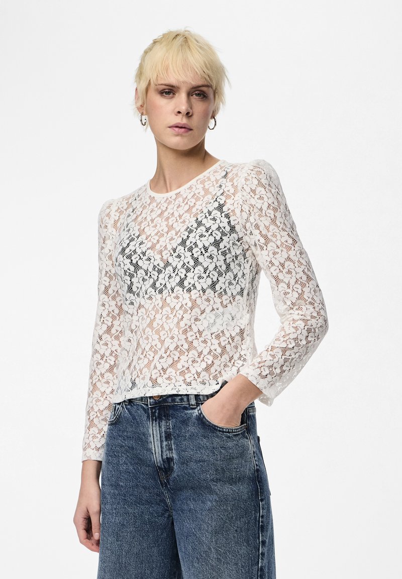 Fehér csipkés, hosszú ujjú felső virágmintákkal, kerek nyakkivágással és cropped hem metszettel, fekete bralette-re rétegezve és farmernadrággal párosítva.