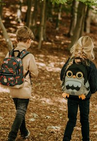 Twee kinderen wandelen door een bos, met rugzakken. De een heeft een donkere tas met vospatronen; de ander heeft een grijze tas in de vorm van een uil met grote ogen.