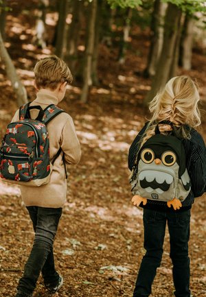 Twee kinderen wandelen door een bos, met rugzakken. De een heeft een donkere tas met vospatronen; de ander heeft een grijze tas in de vorm van een uil met grote ogen.
