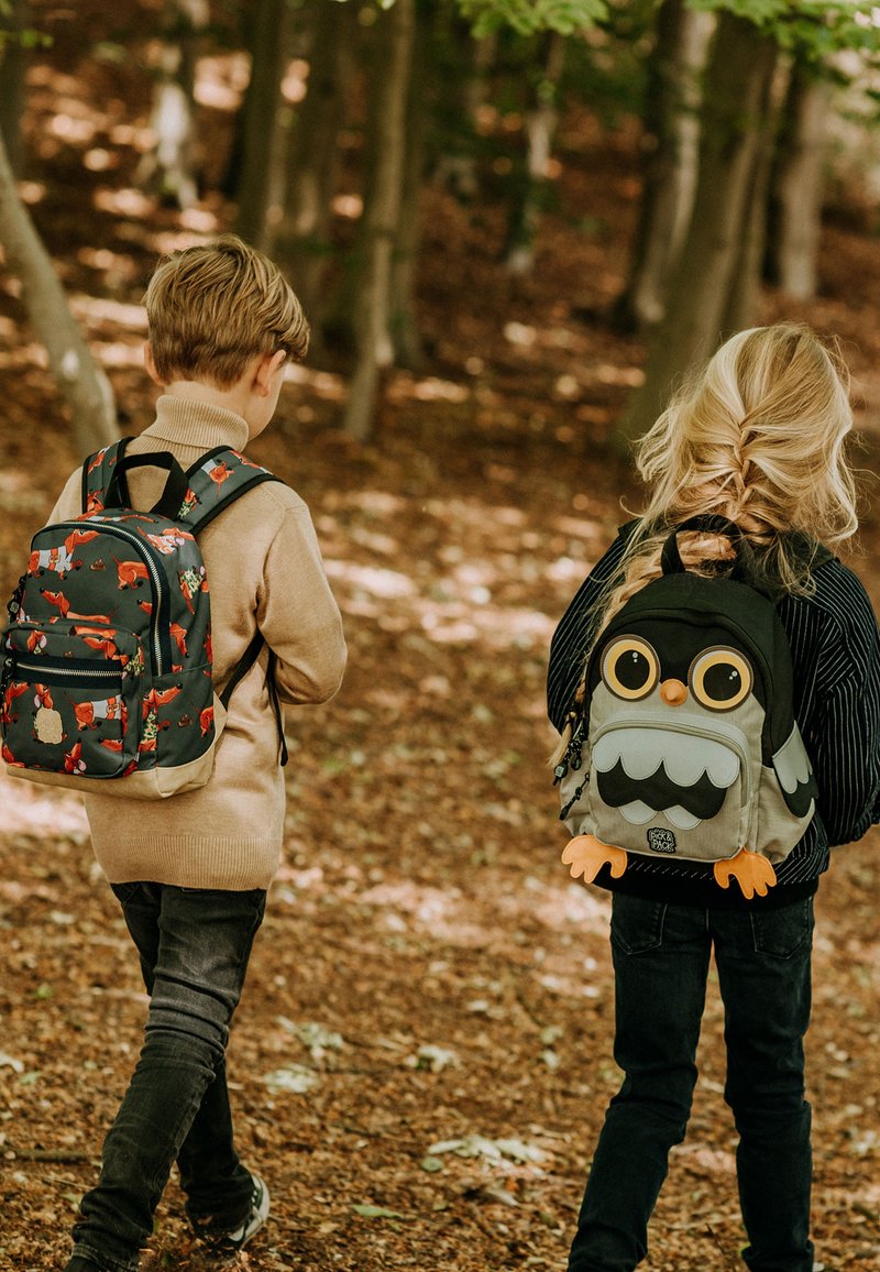 Twee kinderen wandelen door een bos, met rugzakken. De een heeft een donkere tas met vospatronen; de ander heeft een grijze tas in de vorm van een uil met grote ogen.