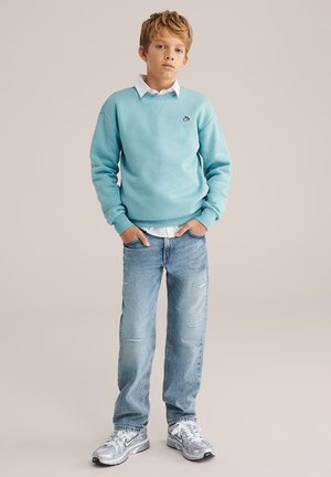 Svetlomodrá mikina s okrúhlym výstrihom v páre s ľahko ošúchanými svetlými džínsami. Oblečenie dopĺňajú sivé tenisky s čiernymi detailmi.