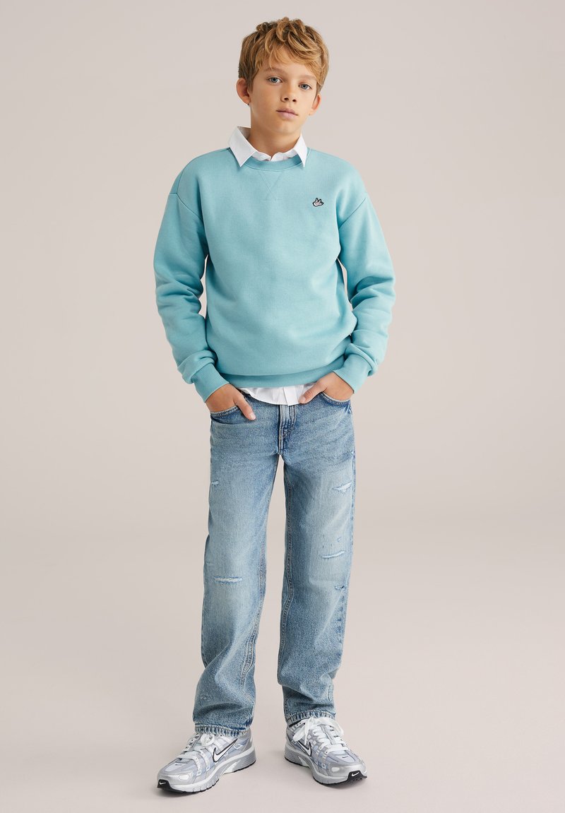 Svetlomodrá mikina s okrúhlym výstrihom v páre s ľahko ošúchanými svetlými džínsami. Oblečenie dopĺňajú sivé tenisky s čiernymi detailmi.