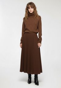 Maglione a collo alto marrone e gonna midi plissettata in marrone coordinato. La gonna presenta plissettature verticali, abbinata a stivali neri con tacco.