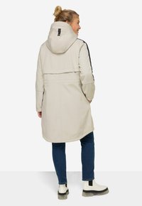Cappotto lungo beige con cappuccio, caratterizzato da accenti neri ai lati, vita stretta e tasche chiuse. Abbinato a jeans scuri e stivali bianchi.