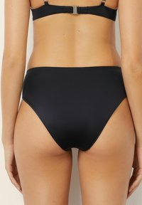 Slip de baie negru cu talie înaltă, cu textură netedă, acoperire minimă și un design elegant. Prezintă o tăietură subtilă pe părțile laterale.