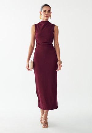 Femme portant une robe midi bordeaux sans manches, des boucles d’oreilles et bracelets dorés, tenant une pochette beige, et portant des talons dorés à brides.