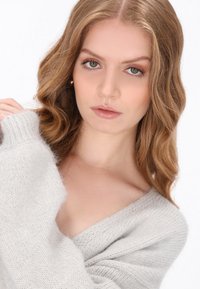Lichtgrijze gebreide trui met een V-hals. Zachte textuur, losse pasvorm en geribbelde manchetten. Model heeft golvend haar en een natuurlijke make-up.