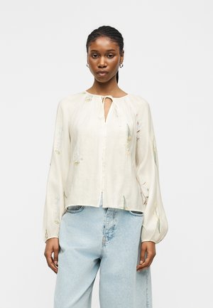 Kobieta nosząca luźną kremową bluzkę z kwiatowymi haftami i długimi bufiastymi rękawami, połączoną z jasnoniebieskimi, wysokim stanem jeansami.