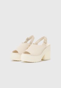 Sandales à plateforme beiges avec un bout ouvert, un design à bride arrière et un talon épais. Fabriquées en tissu doux avec une semelle texturée et des accents dorés.