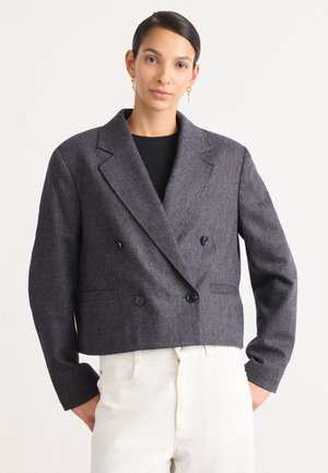 Abgeschnittenes, doppelt geknöpftes Blazer in dunkelgrauem Fischgrätenstoff mit zwei Knöpfen, Reverskragen und Seitentaschen. Kombiniert mit hellen Hosen.