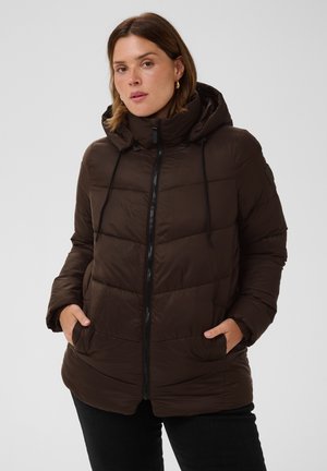 Mujer con chaqueta acolchada con capucha de color marrón oscuro, cremallera frontal y manos en los bolsillos, de pie frente a un fondo neutro.