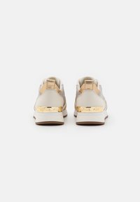 Paire de baskets femme beige et or avec plaque logo dorée sur semelles compensées blanches, vue de dos sur fond blanc.