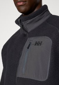 Fleecejacke in Dunkelgrau mit hohem Kragen, halblangem Reißverschluss an der Vorderseite und einer Brusttasche mit blauem Reißverschluss und Logo. Strukturierter Stoff.