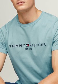 Světle modré bavlněné tričko s námořnickým logem "TOMMY HILFIGER" s červeným a bílým detailem a textem "EST. 1985" pod ním. Krátké rukávy a kulatý výstřih.