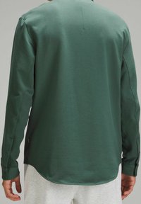 Camisa de manga larga, verde oscuro, hecha de una tela suave, con dobladillo redondeado, puños abotonados y un diseño sencillo.