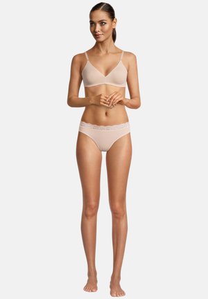 Frau steht barfuß, trägt einen hautfarbenen BH und Spitzenhöschen, schaut leicht zur Seite und hat die Hände verschränkt.