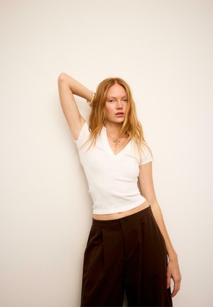 Femme aux longs cheveux blonds portant un haut blanc à manches courtes et un pantalon marron foncé, posant avec un bras levé contre un mur beige uni.