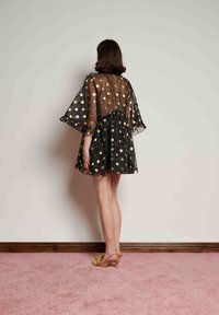 Μαύρο φόρεμα με πουά και χρυσές λεπτομέρειες, διάφανο υπερκείμενο ύφασμα, μανίκια με βολάν και φουσκωτή φούστα. Το μοντέλο φοράει ανοιχτά τακούνια σε χρώμα ταμπά, φωτογραφημένο από πίσω.