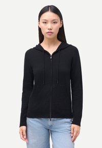 Svart huvtröja med dragkedja och huva, gjord av mjukt tyg, med framfickor och dragsnören på huvan, kombinerad med ljusblå jeans.