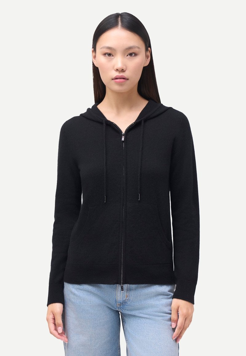 Svart huvtröja med dragkedja och huva, gjord av mjukt tyg, med framfickor och dragsnören på huvan, kombinerad med ljusblå jeans.