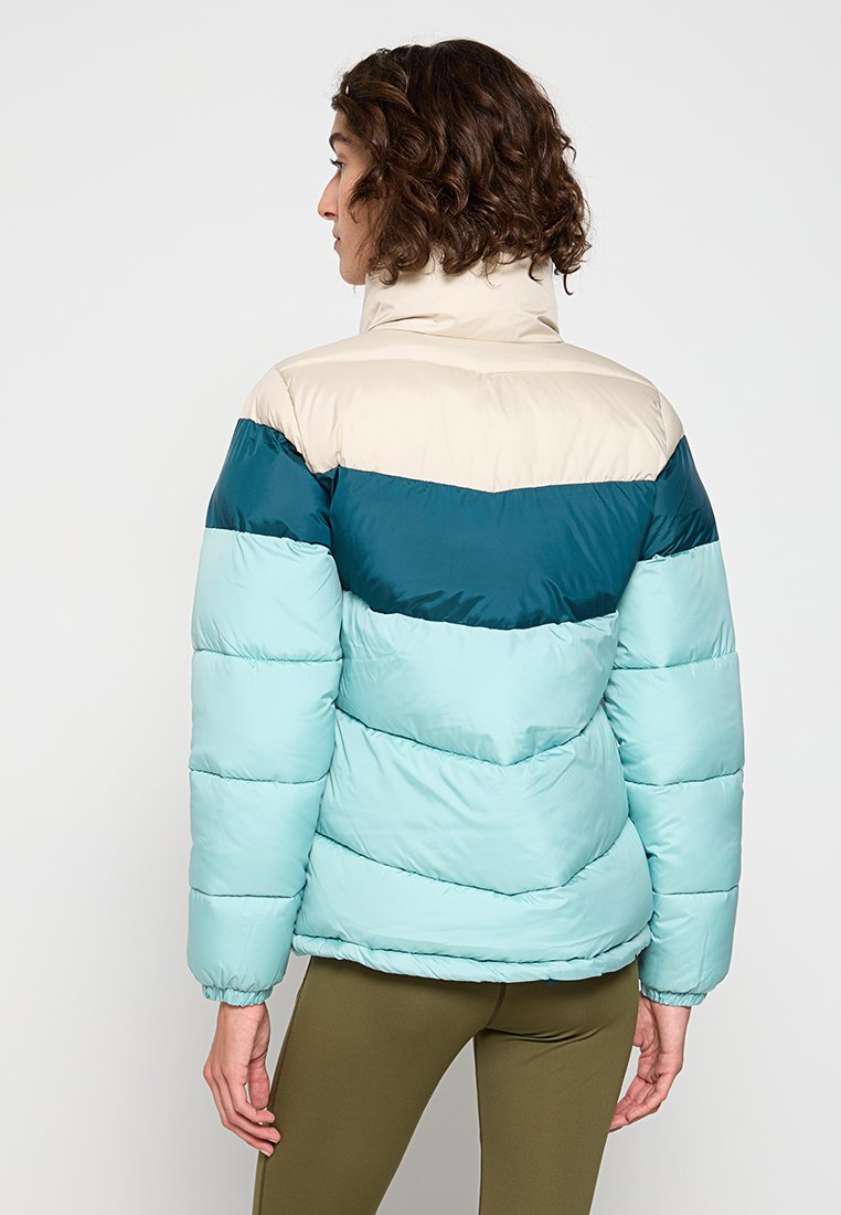 Mujer con cabello rizado corto que lleva una chaqueta acolchada de tres tonos en beige, verde azulado oscuro y azul claro, de pie de espaldas.