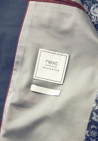 Hallitus halli kangast riietus marooni aktsentide ja paremal küljel oleva mustriga, millel on valge silt "next TAILORING" ja kangassilmus.