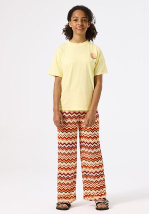 Geel katoenen t-shirt met een kleine graphic op de borst, gecombineerd met brede broek met een rood en geel zigzagpatroon en zwarte sandalen.