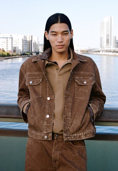 Young person with long dark hair wearing a brown jacket and matching pants stands by a river railing with city buildings in the background.