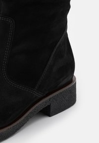 Bottine noire en daim avec surpiqûres visibles et talon bloc en caoutchouc texturé sur fond blanc.