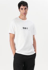 Vit bomull t-shirt med rund halsringning, prydd med svarta grafiska sport symboler och texten "Försvarar sedan 1974" i mitten.