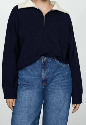 Personne portant un pull bleu marine à manches longues avec un col demi-fermeture éclair blanc, rentré dans un jean taille haute bleu, sur un fond uni.