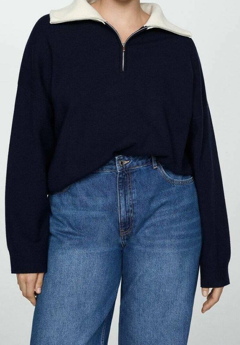Personne portant un pull bleu marine à manches longues avec un col demi-fermeture éclair blanc, rentré dans un jean taille haute bleu, sur un fond uni.