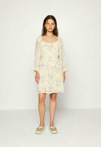 Mulher de pé contra um fundo branco, vestindo um vestido longo de manga comprida com padrão floral em tons pastel e sandálias de plataforma verde claro.