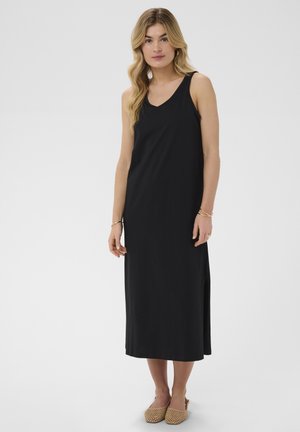 Pikade blondide juustega naine, kes kannab varrukateta musta midi-kleiti ja beežist punutud slip-on jalanõusid, seisab tavalise tausta ees.