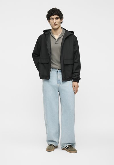 Άνδρας με σγουρά μαλλιά, φορώντας μαύρο μπουφάν με κουκούλα, γκρι πόλο πουκάμισο, φαρδύ ανοιχτό μπλε τζιν και πράσινα αθλητικά παπούτσια, στέκεται μπροστά σε λευκό φόντο.