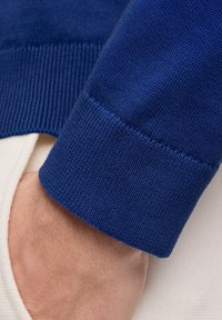 Blauer gestrickter Pullover mit geripptem Bündchen, kombiniert mit einem weißen Hemd darunter, das die kontrastierenden Farben und Materialien hervorhebt.