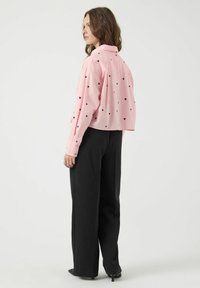 Chemise rose courte avec des motifs de cœurs noirs, manches longues, poignets à boutons, associée à un pantalon large noir et des talons noirs.
