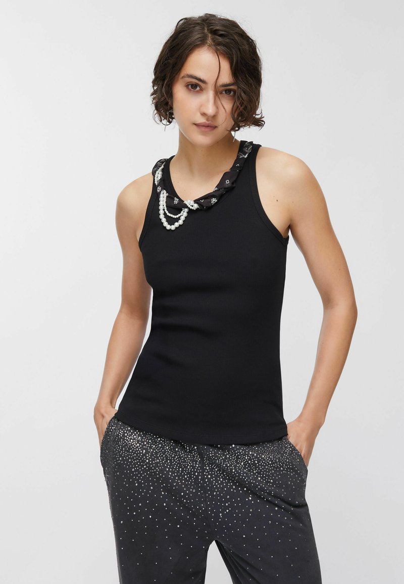 Débardeur noir à texture côtelée, avec un col noir à motif orné de trois rangs de perles blanches. Porté avec un pantalon noir scintillant.