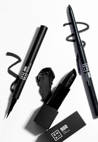 Svart leppestift i et rektangulært etui, en svart eyelinerpenn med spiss tupp, og en svart leppepenn, alt arrangert med en svart virvel.